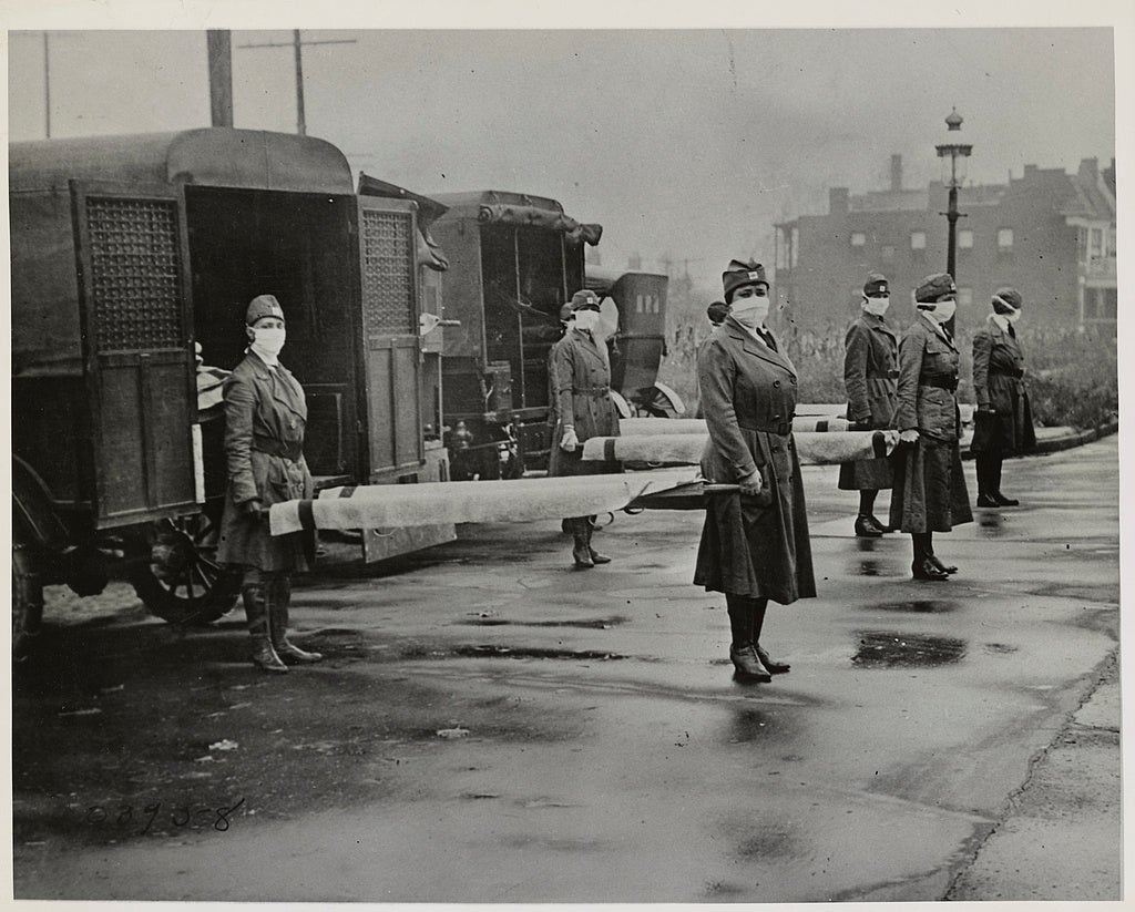 In preparation for the deadly flu, mask-wearing women hold stretchers at backs of ambulances.